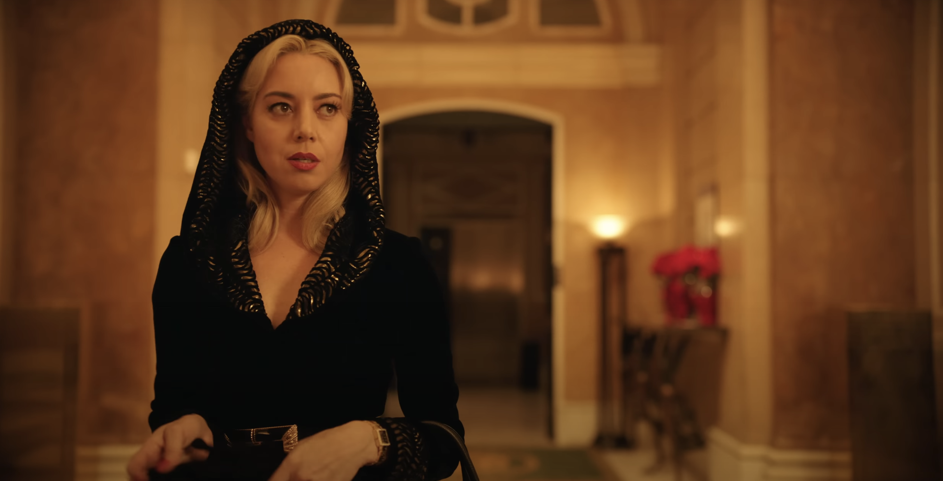 Aubrey Plaza in a scene from a movie, wearing a hooded garment with gold accents, inside an ornate building lobby