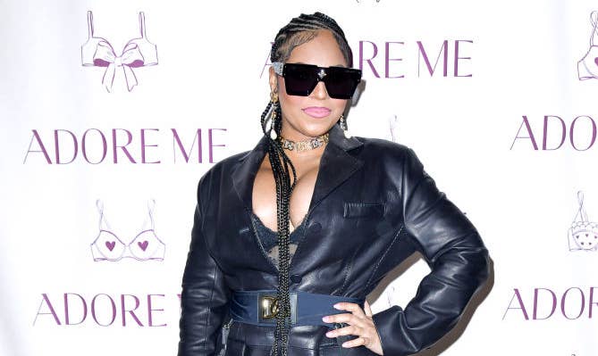 Ashanti stands on the red carpet in a black leather blazer with fishnet stockings, sunglasses, and braided hair, in front of an "Adore Me" step and repeat banner