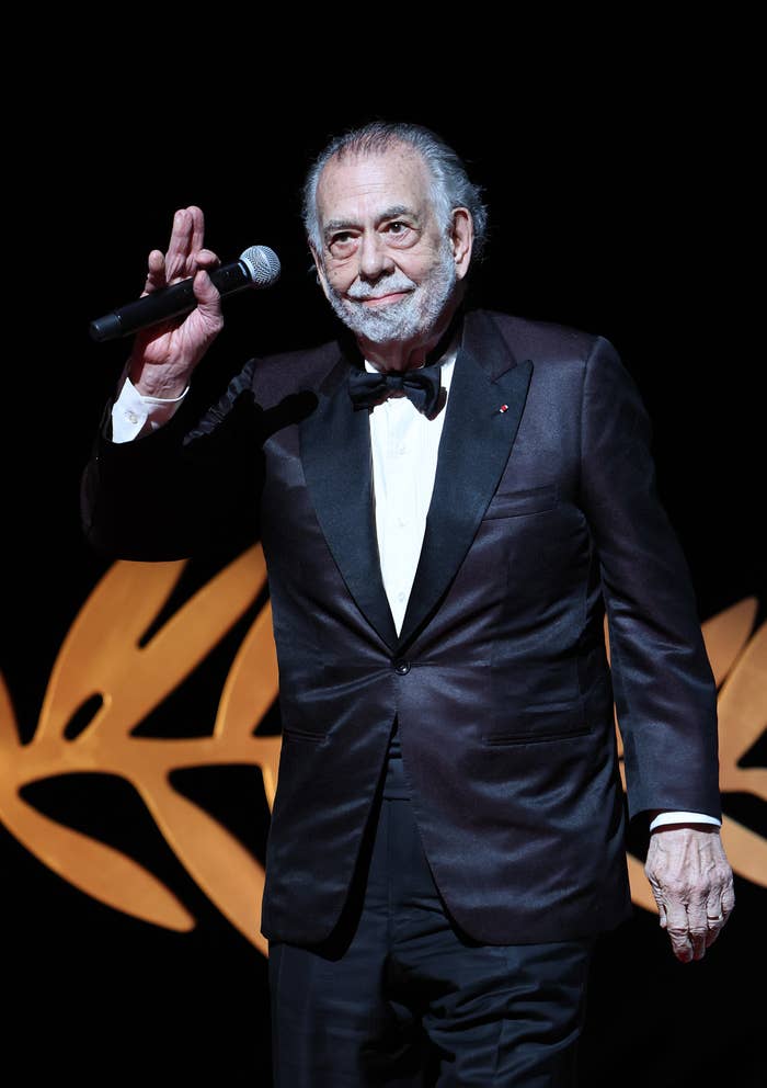Francis Ford Coppola, in formal attire with a bow tie, waves while holding a microphone at a red carpet event