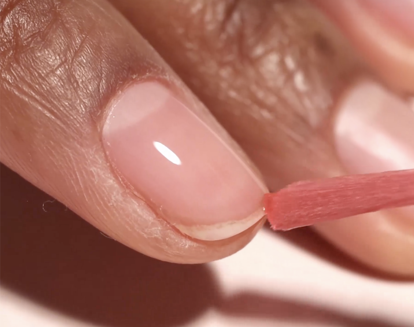 A close-up of a person applying nail polish to their fingernail with a brush. The polished nail shows a glossy finish. The person is unidentified