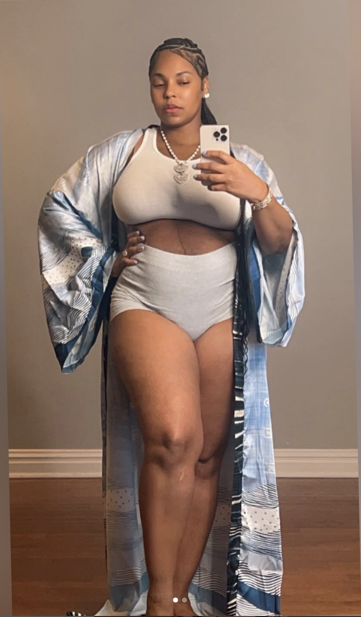 Ashanti stands for a mirror selfie wearing a printed robe, a white crop top, and grey high-waisted shorts. They are holding a phone in one hand and have a relaxed expression