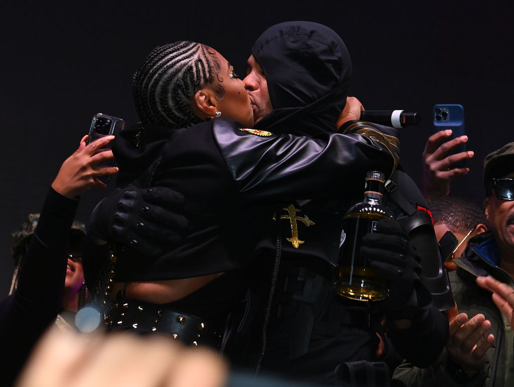 Ashanti and Nelly share a kiss on stage while holding phones and bottles, surrounded by fans and photographers