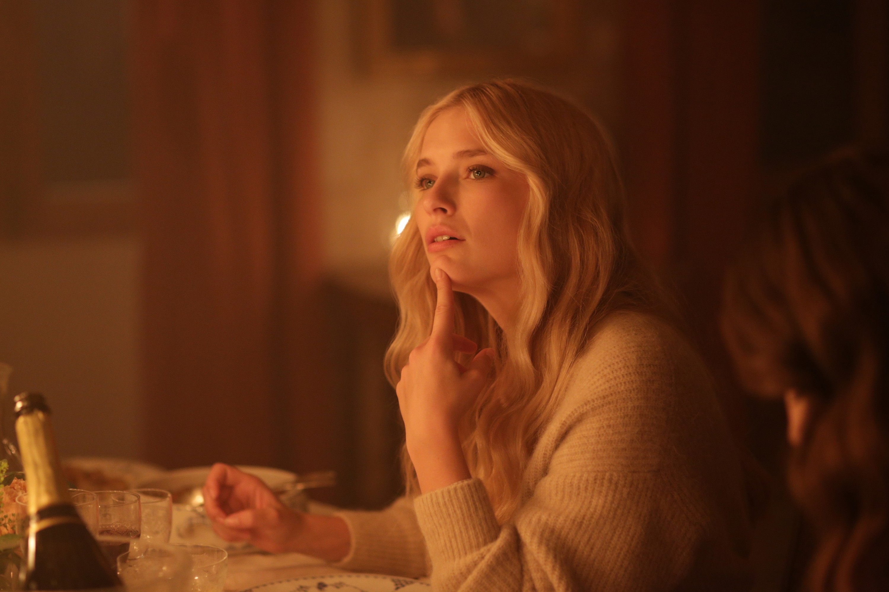 Woman with long hair wearing a sweater sits at a table with a thoughtful expression, looking upward