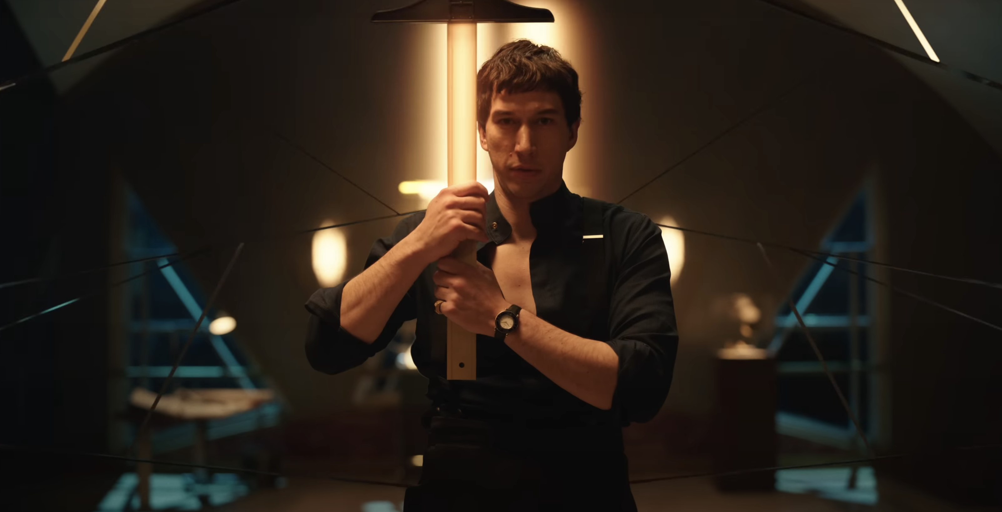 Adam Driver is holding an illuminated sword-like object in a dimly lit room with modern furnishings, giving an intense look