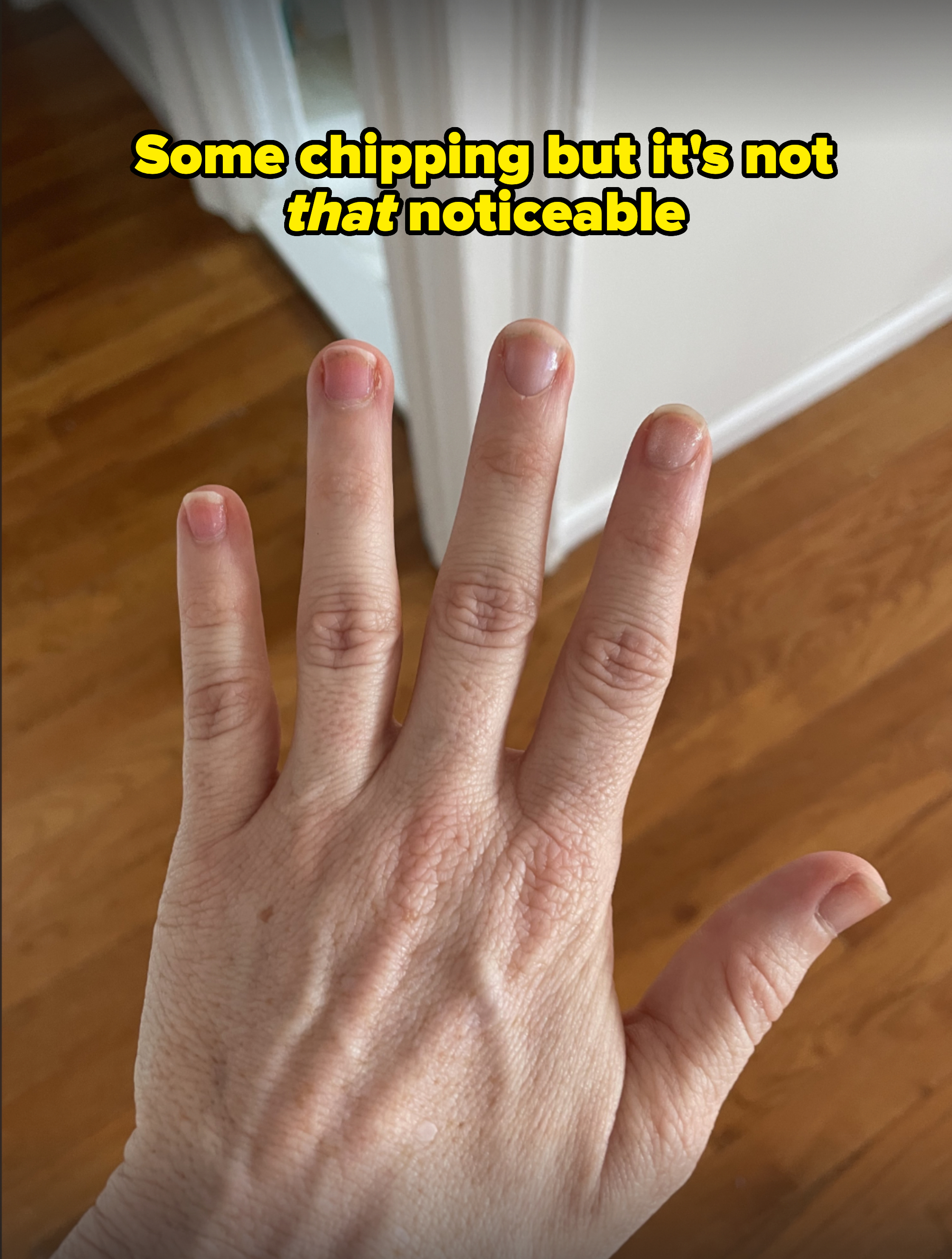 A close-up of a person's left hand with short, unpolished nails against a wooden floor background