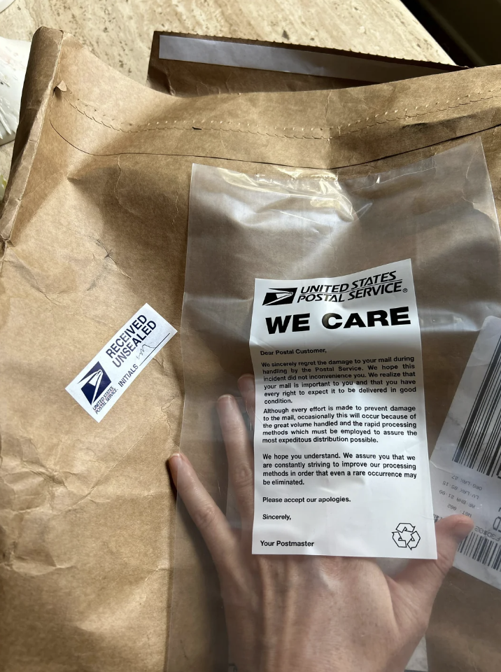 A person holding a "We Care" notice from the United States Postal Service, indicating that their package was damaged during processing. There is another envelope in the background