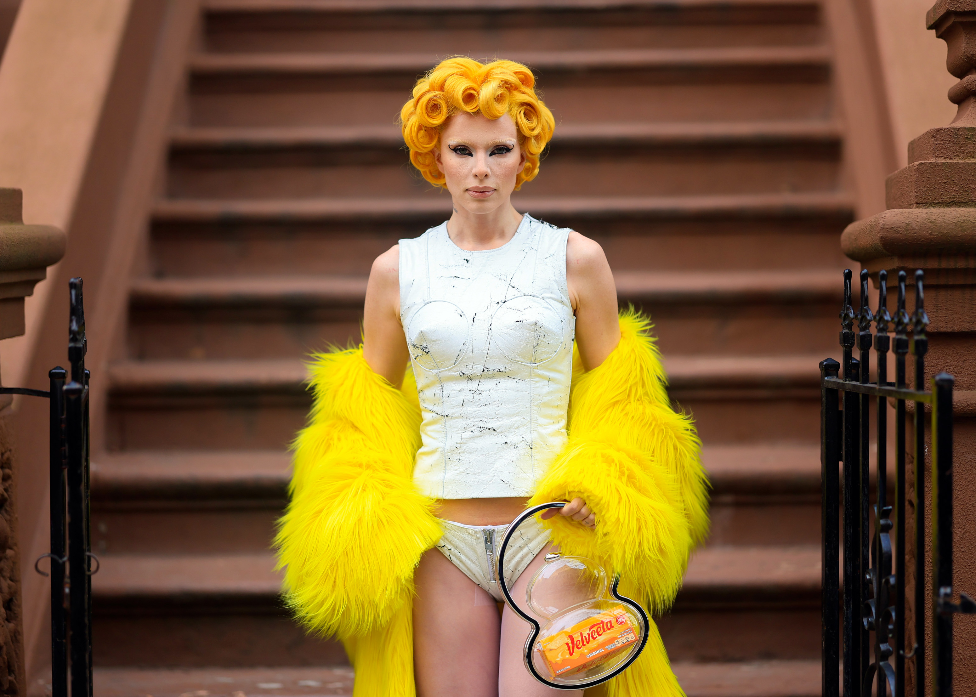 Julia Fox stands on steps in a white sleeveless top, yellow feathered coat, and silver underwear, holding a clear heart-shaped purse labeled "Italian."
