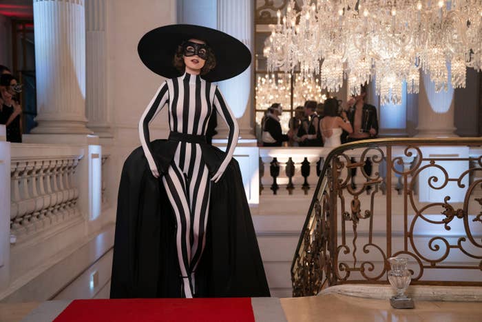 Lily Collins stands on a red carpet in an elegant hallway, wearing a bold black and white striped gown, paired with a wide-brimmed hat and a black mask