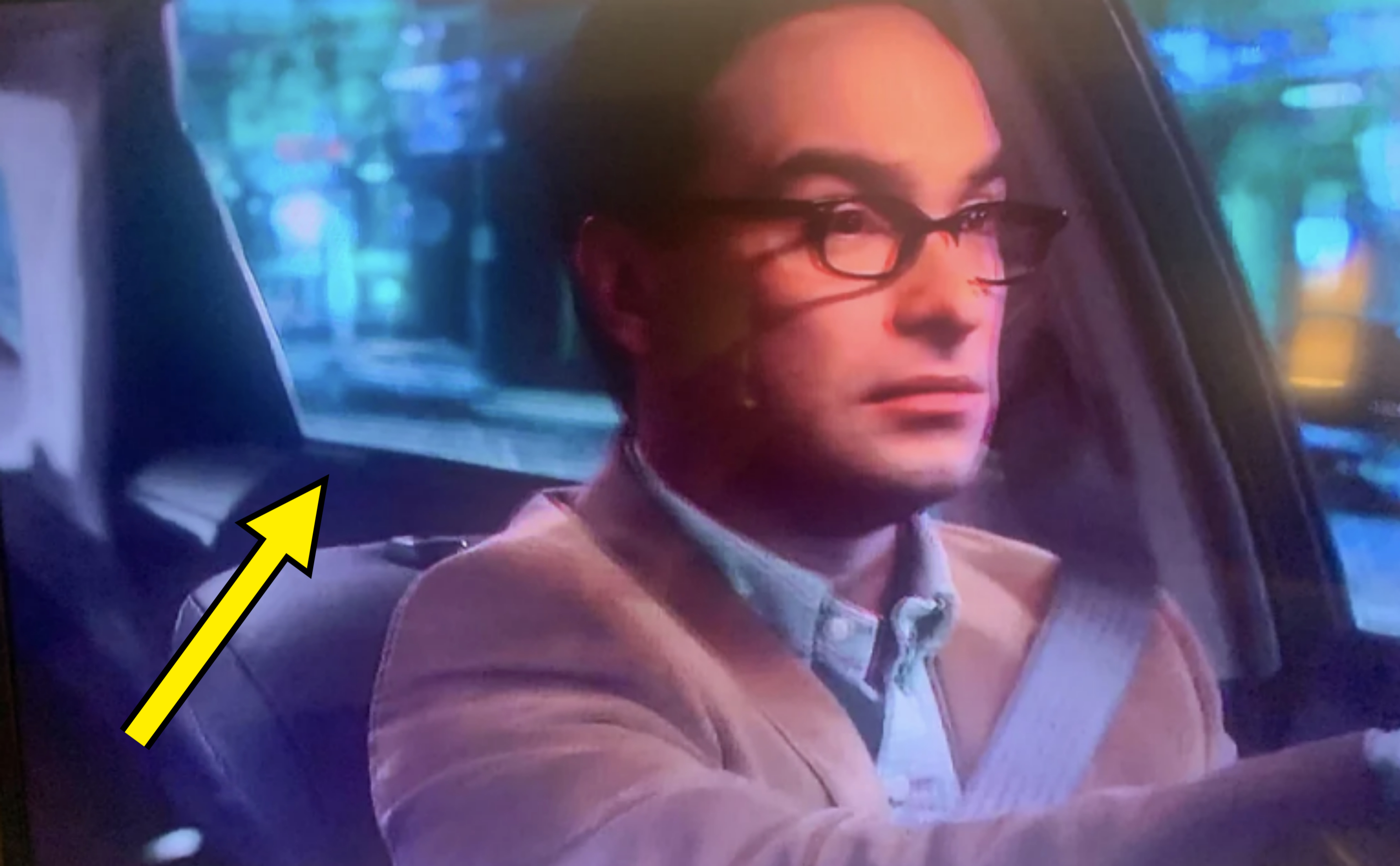 Johnny Galecki, wearing glasses and a beige jacket over a button-down shirt, is driving a car at night. The background shows blurred city lights