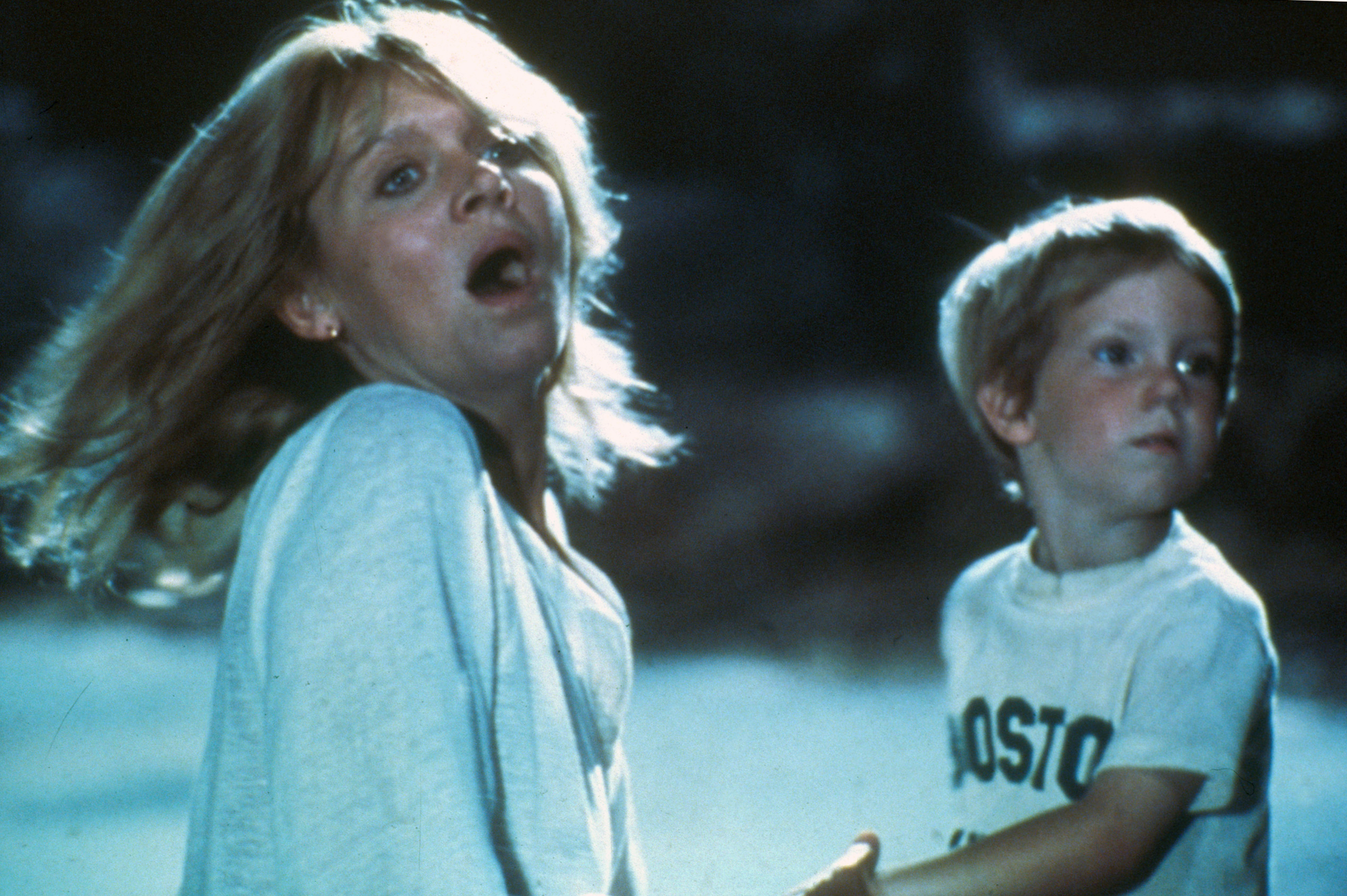 A woman with long hair holds a young boy wearing a "Boston" t-shirt, both looking startled or scared