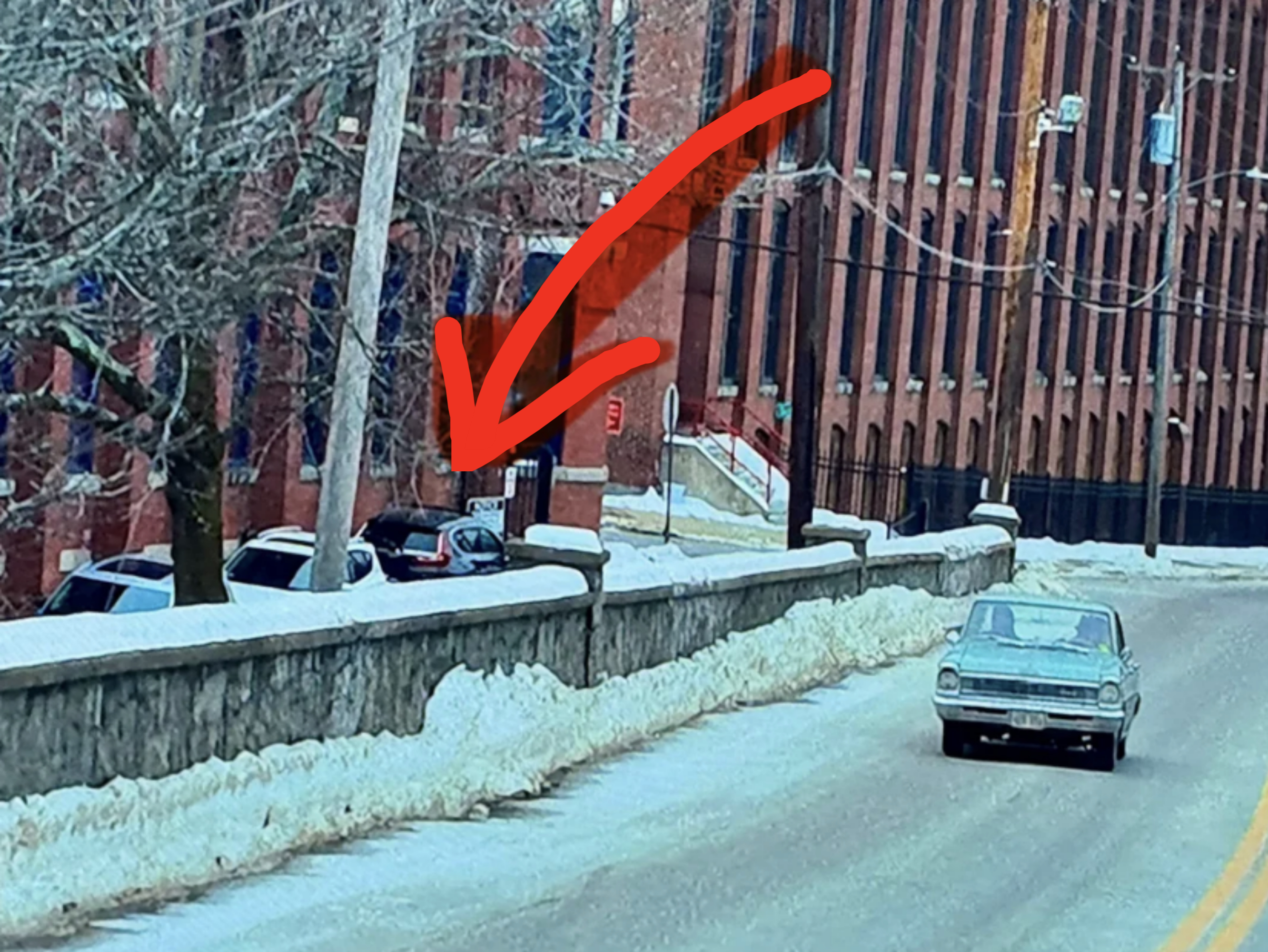 A snowy street with a gray vintage car driving by a row of parked cars near a brick building. An orange arrow points towards the building