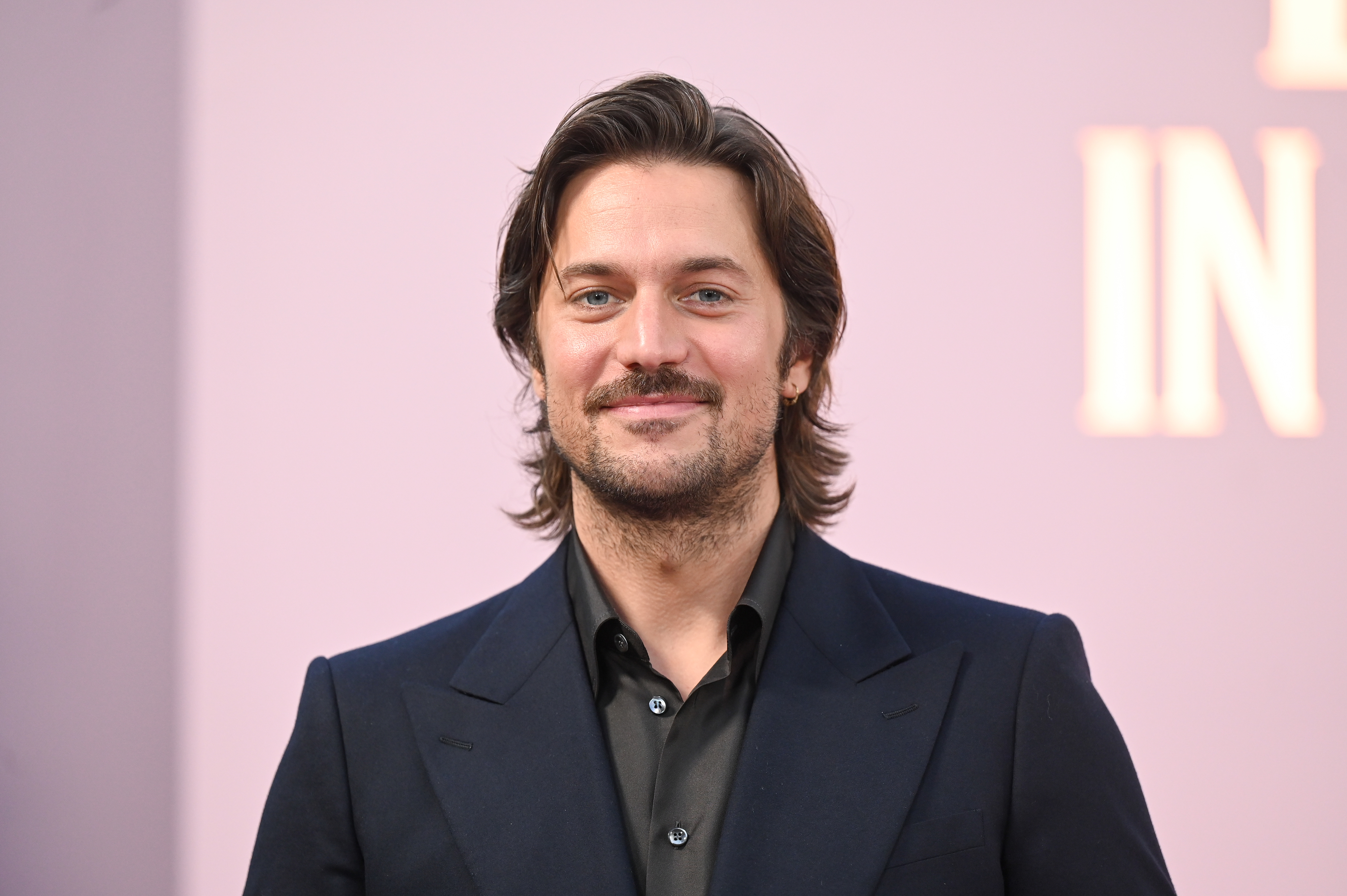 A man with shoulder-length hair and a beard wearing a black suit and black shirt, smiling at a red carpet event