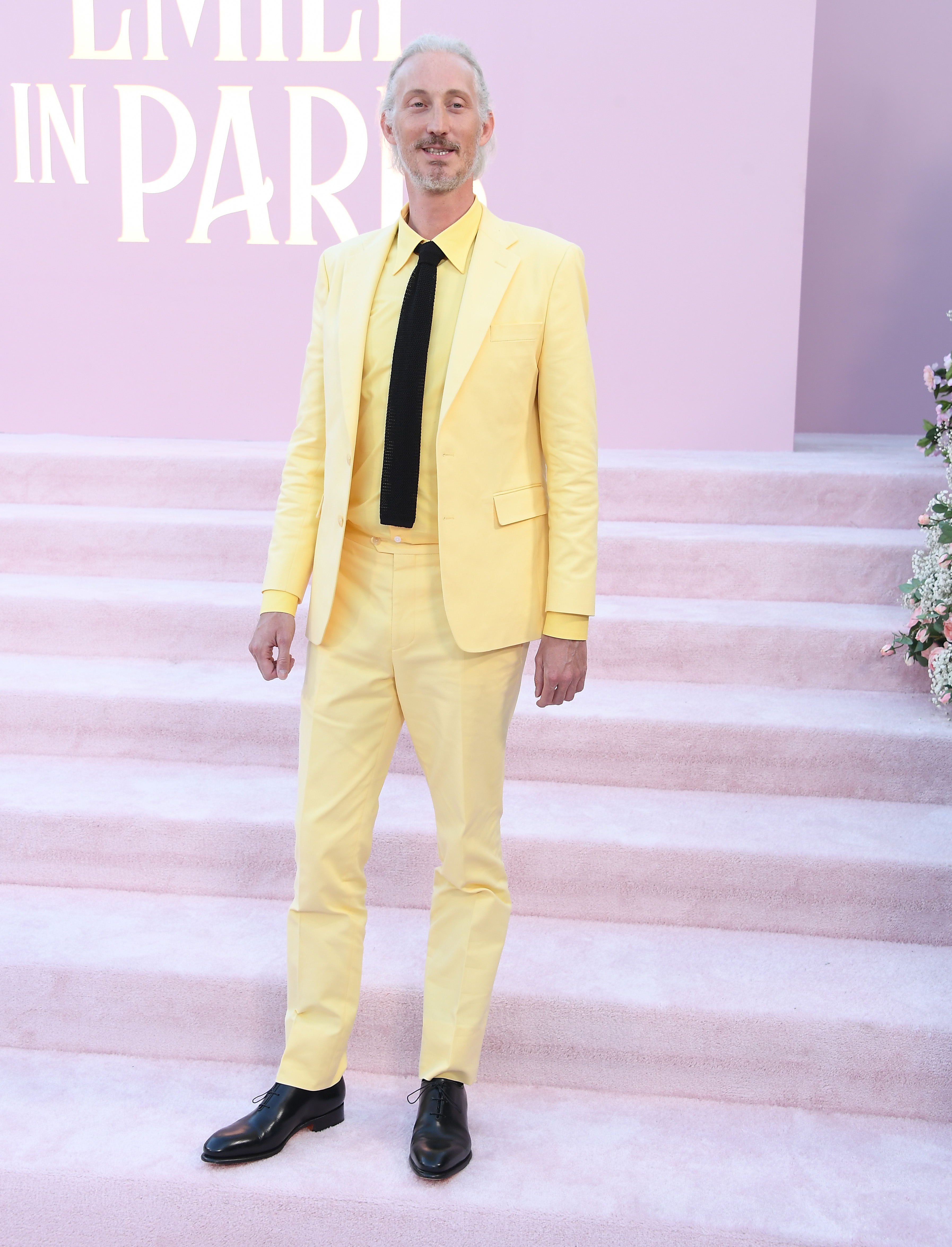 A man in a yellow suit with a black tie stands on steps at an "Emily in Paris" event
