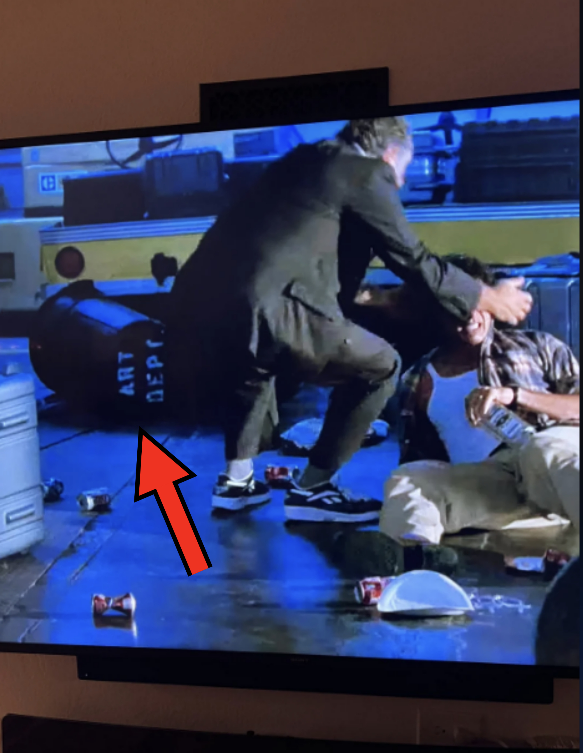 A man dressed in dark clothing and sneakers aggressively grabs another man sitting on the ground and holding a bottle, against a backdrop of scattered items