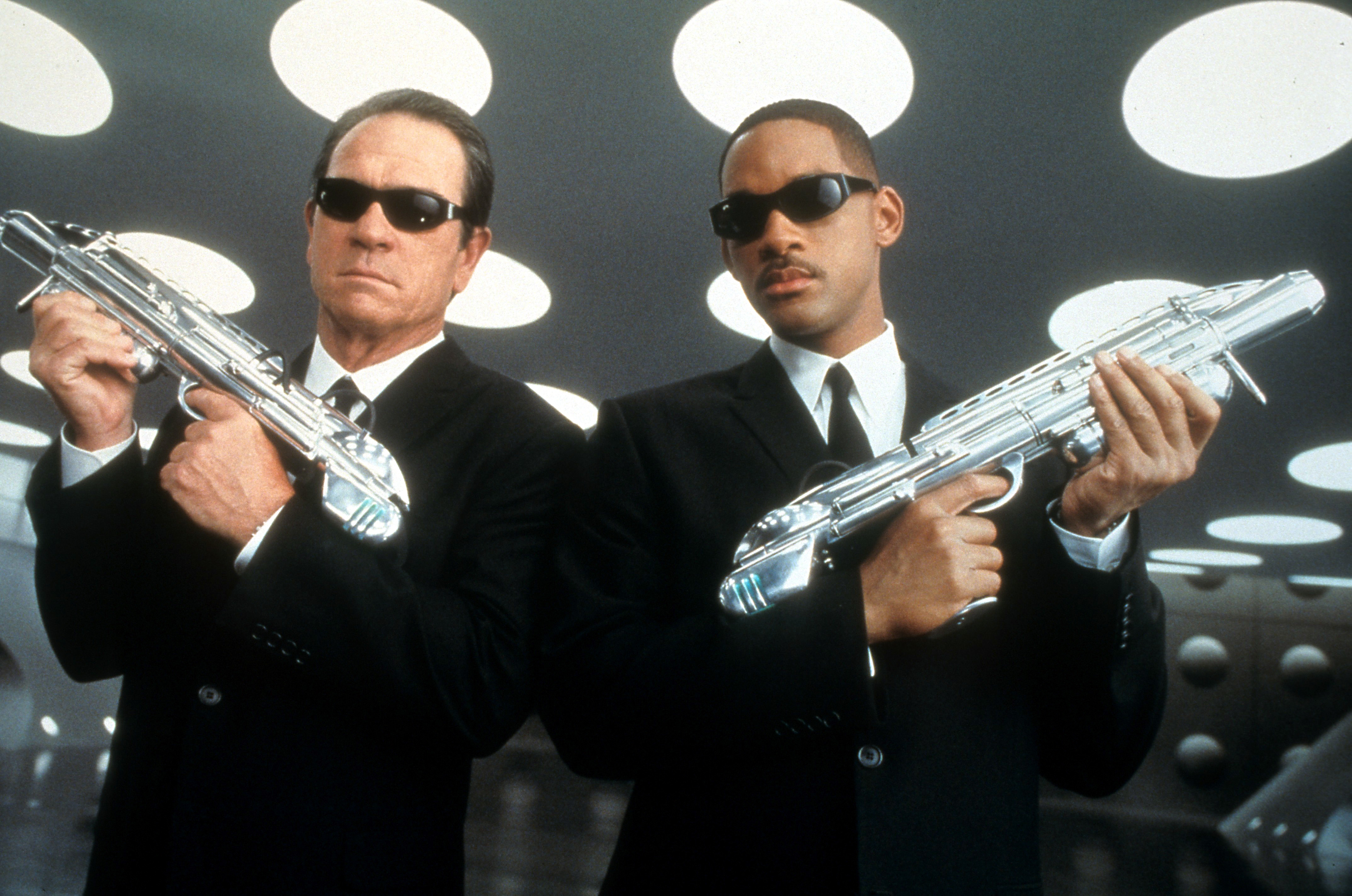 Tommy Lee Jones and Will Smith, dressed in black suits and sunglasses, holding futuristic weapons against a backdrop with circular lights