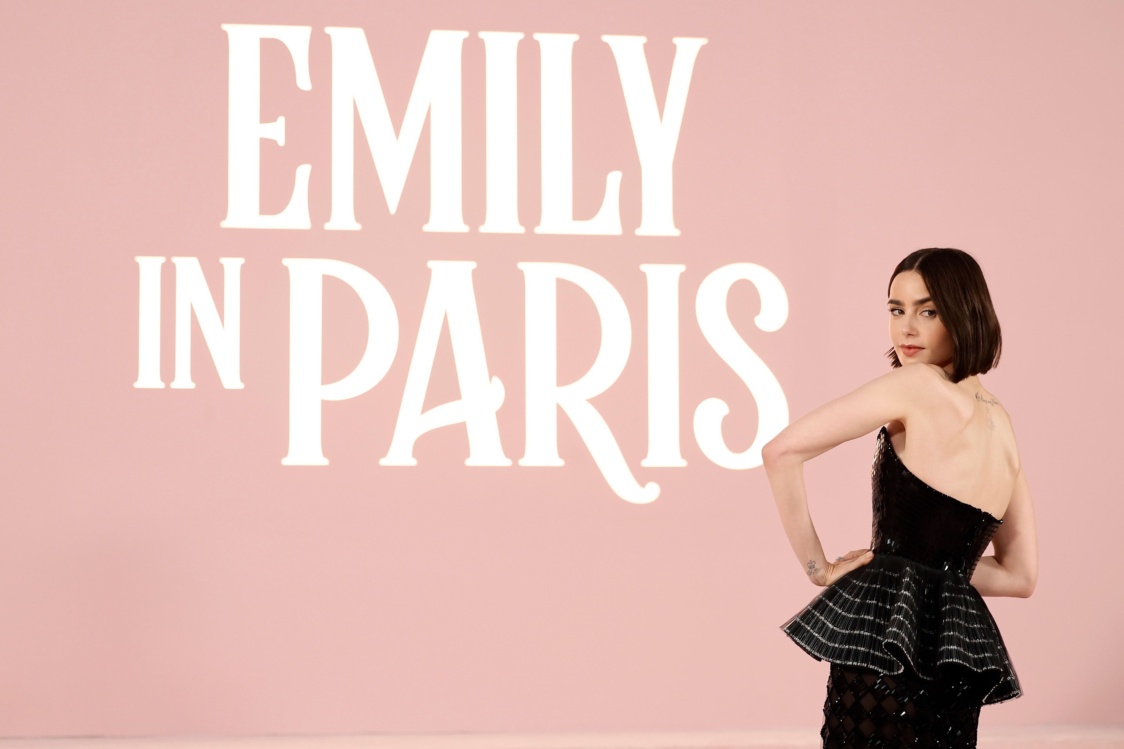 Lily Collins, in an elegant dress, poses in front of a backdrop with text "Emily in Paris"