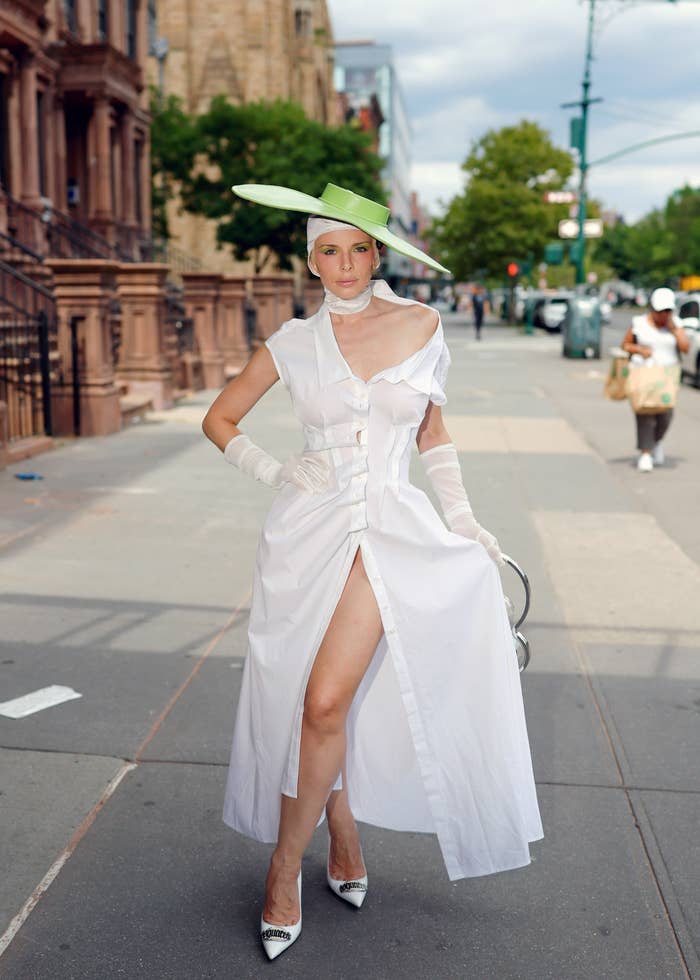 Julia Fox poses outdoors in a stylish vintage-inspired dress with a high slit, matching gloves, and a large hat. She is standing on a city sidewalk