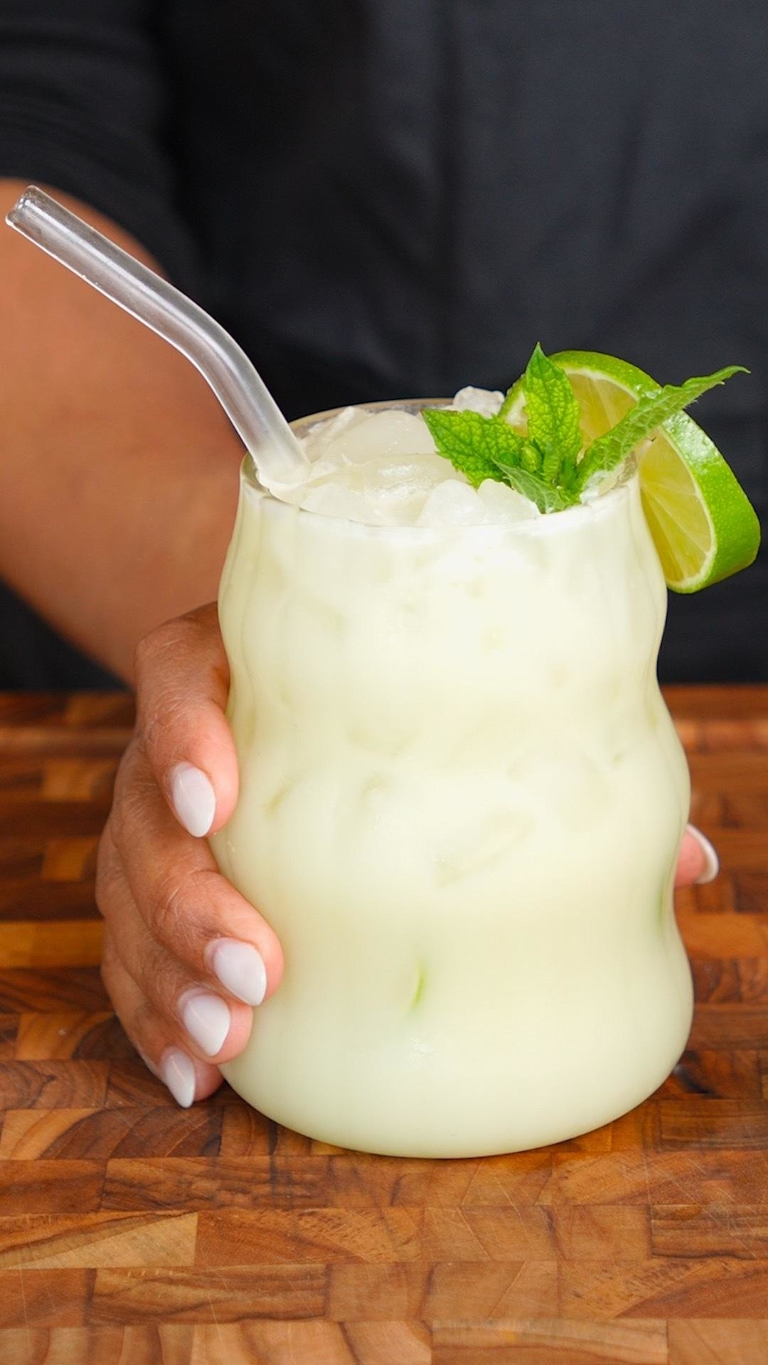 A hand holding a frothy, creamy drink with ice in a textured glass, garnished with a lime wedge and mint sprig on a wooden surface