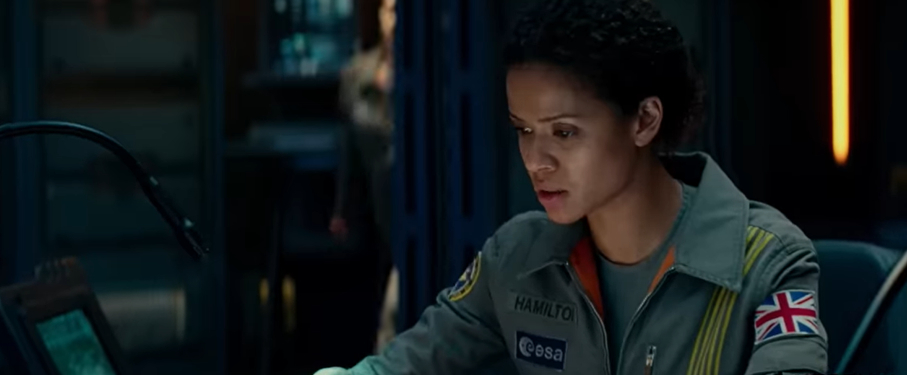 Gugu Mbatha-Raw, as Hamilton, in a space suit with the ESA and UK flag patches, seated and focused, in a space station from a scene in "The Cloverfield Paradox."