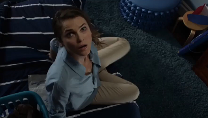 Keri Russell sits on a bed, looking up, in what appears to be a bedroom. She wears a light blue shirt and beige pants