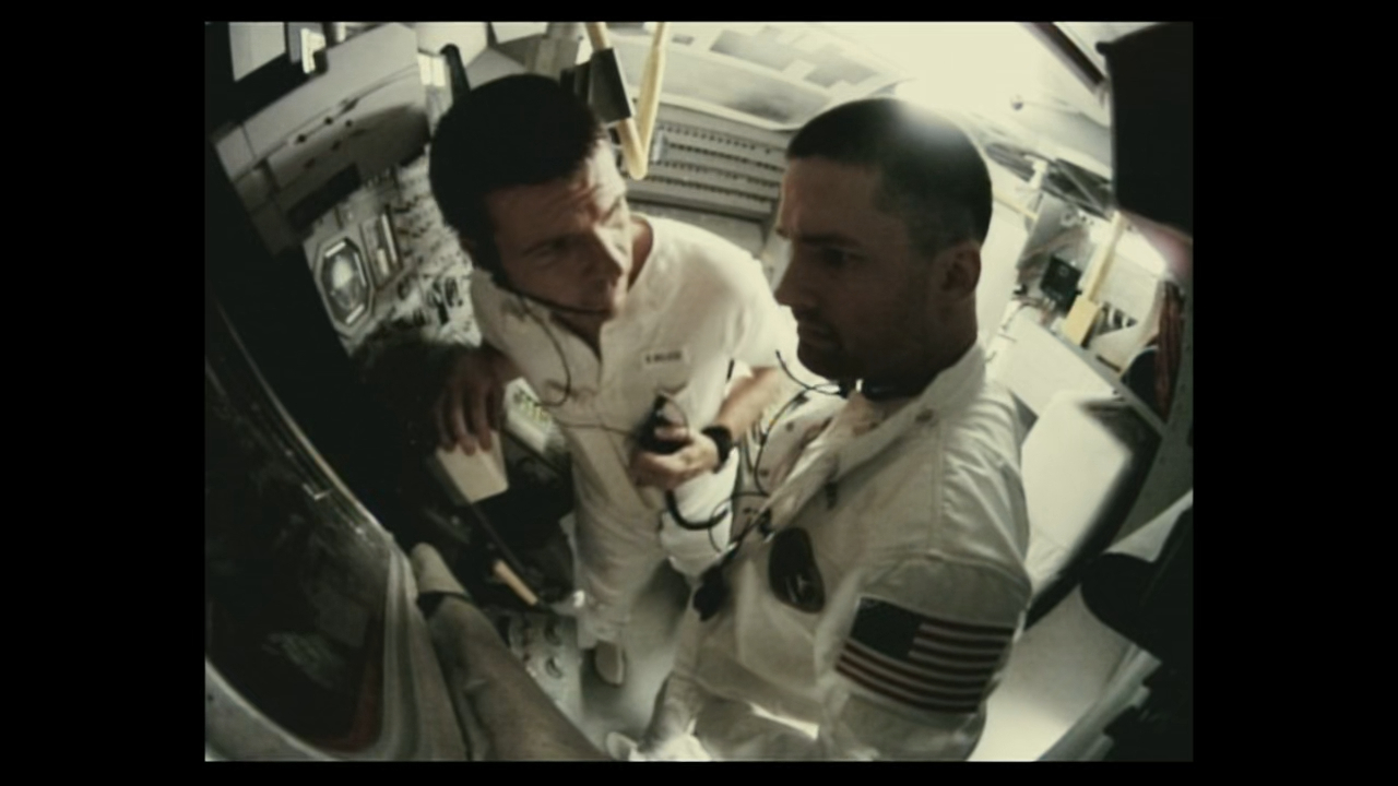 In a scene from Apollo 18, two astronauts, one holding a communication device, collaborate on a task inside a spacecraft. Their suits show NASA emblems and the American flag