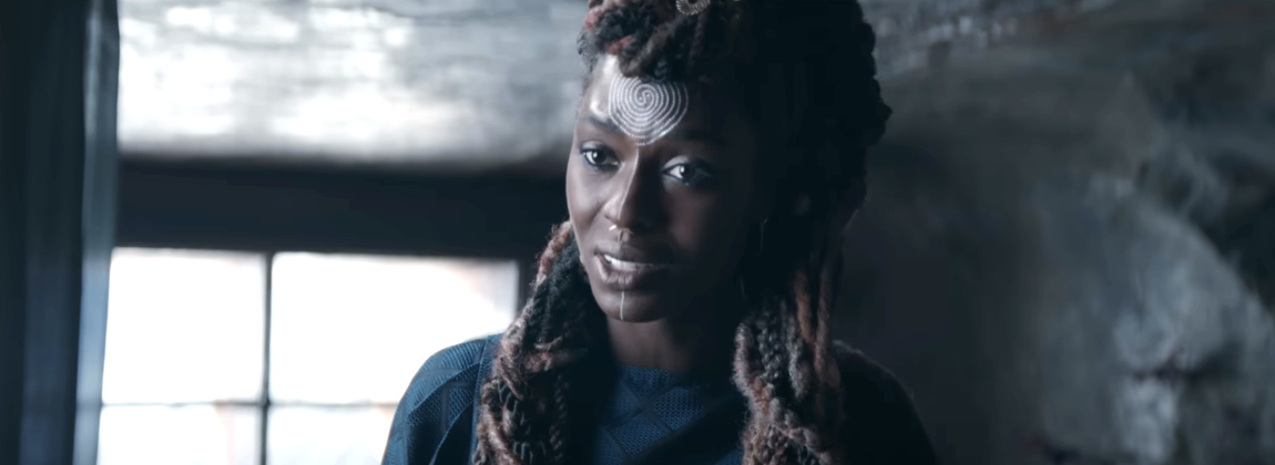 Person with intricate face paint and dreadlocks in an indoor, slightly dim setting, wearing a dark top