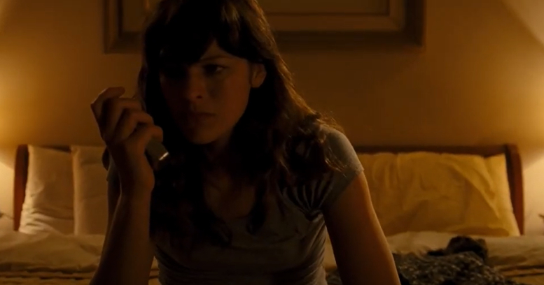 Milla Jovovich with long wavy hair sits on a bed in a dimly lit room, holding a phone to her ear, looking concerned. No identification is provided for the person