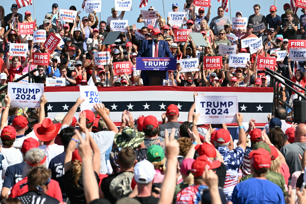 Donald Trump at a rally podium with a crowd holding "Trump 2024" and "Fire Biden" signs