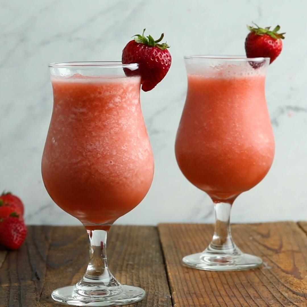 Two glasses of strawberry daiquiris garnished with whole strawberries, sitting on a wooden table