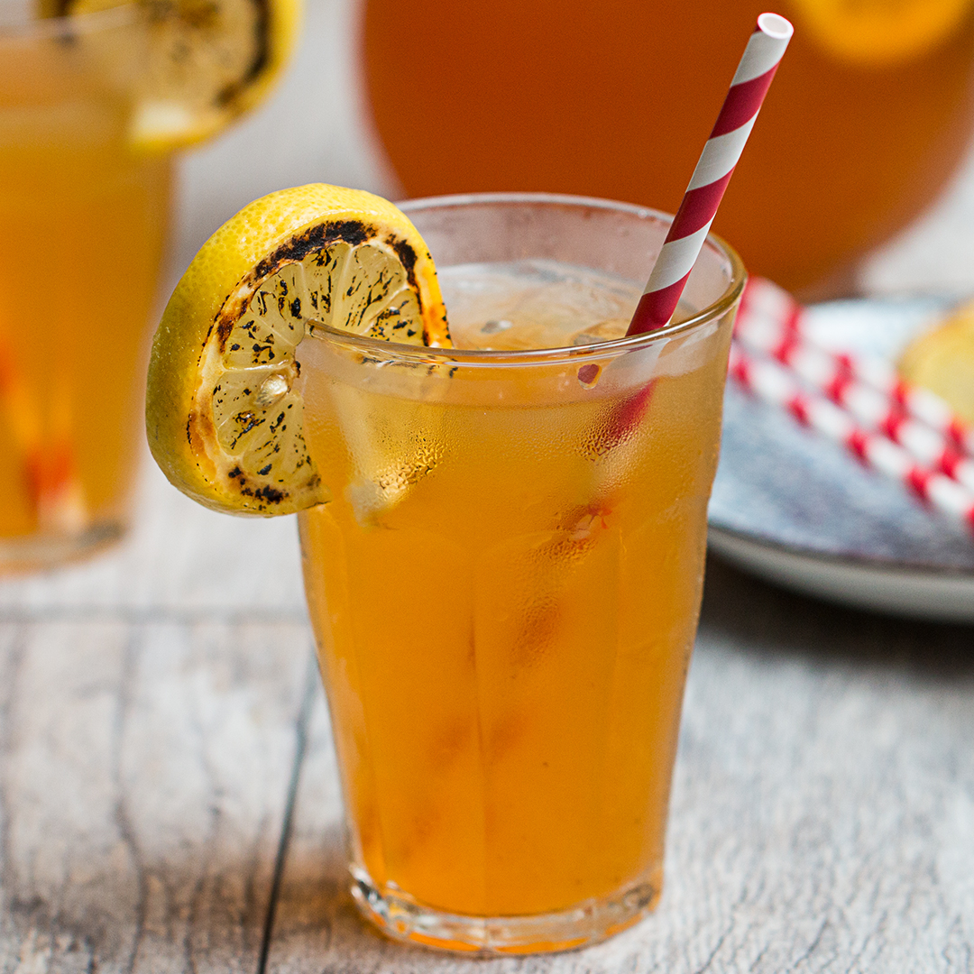 A glass filled with iced tea or lemonade, garnished with a grilled lemon slice and a red and white striped straw. Another glass and plate are in the background