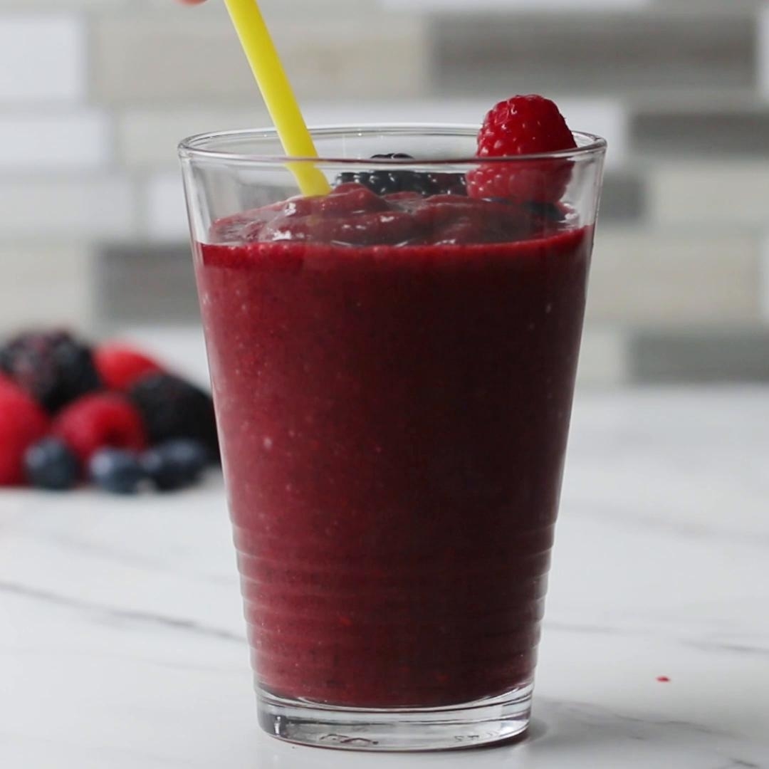 A glass of berry smoothie filled to the rim, garnished with a raspberry on top, and a yellow straw. Fresh berries are scattered in the background