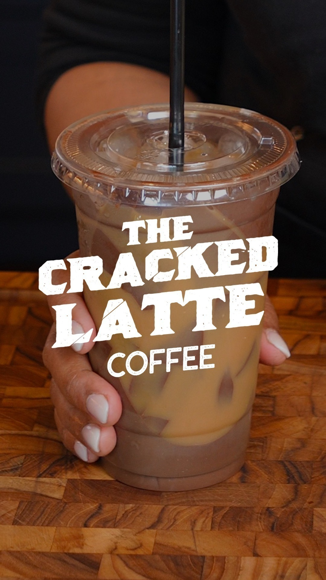A hand holds a plastic cup of iced coffee with a straw. The text on the image reads, &quot;The Cracked Latte Coffee.&quot;
