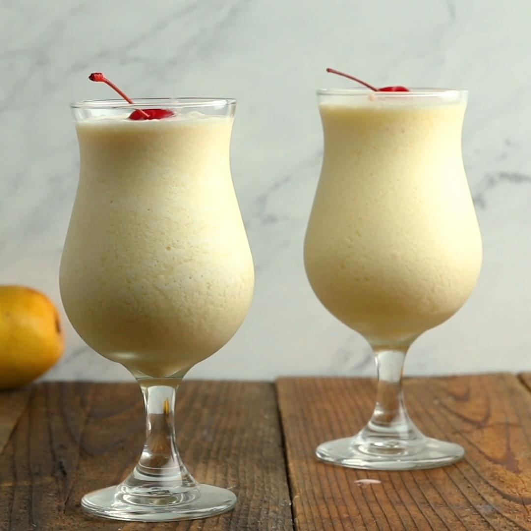 Two tall glasses of creamy yellow frozen mango cocktails, each topped with a cherry, are on a wooden table with a blurred background