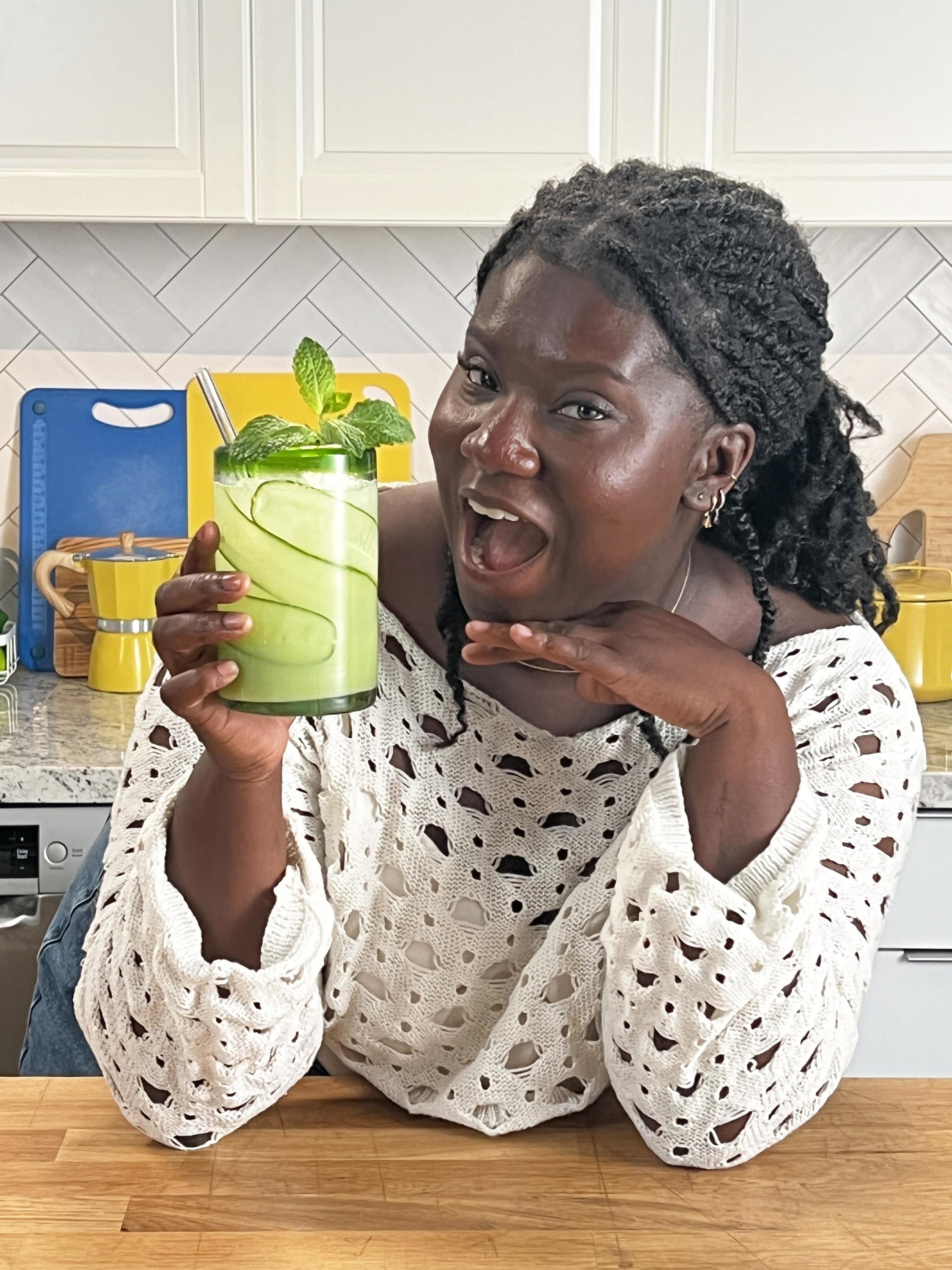 Person in a kitchen holding a green smoothie garnished with mint leaves, smiling excitedly