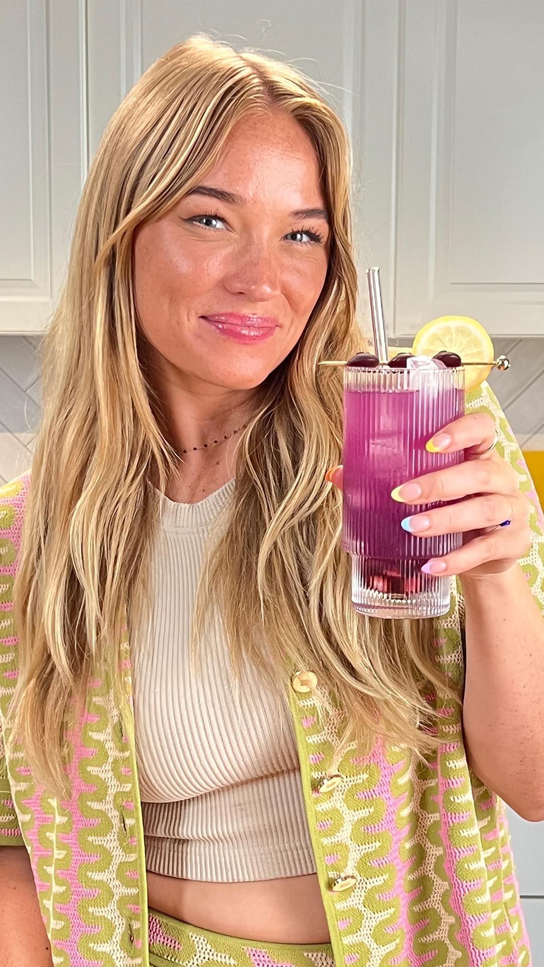Person holding a purple drink garnished with lemon and berries, standing in a kitchen and smiling