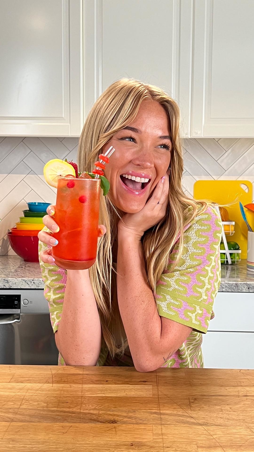 Person smiling in a kitchen, holding a tall glass filled with a fruity red beverage garnished with a lemon slice and berries