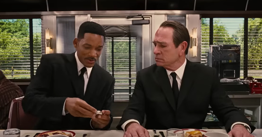 Will Smith and Tommy Lee Jones sit in a diner booth, wearing black suits and white shirts, discussing something while looking at a small device