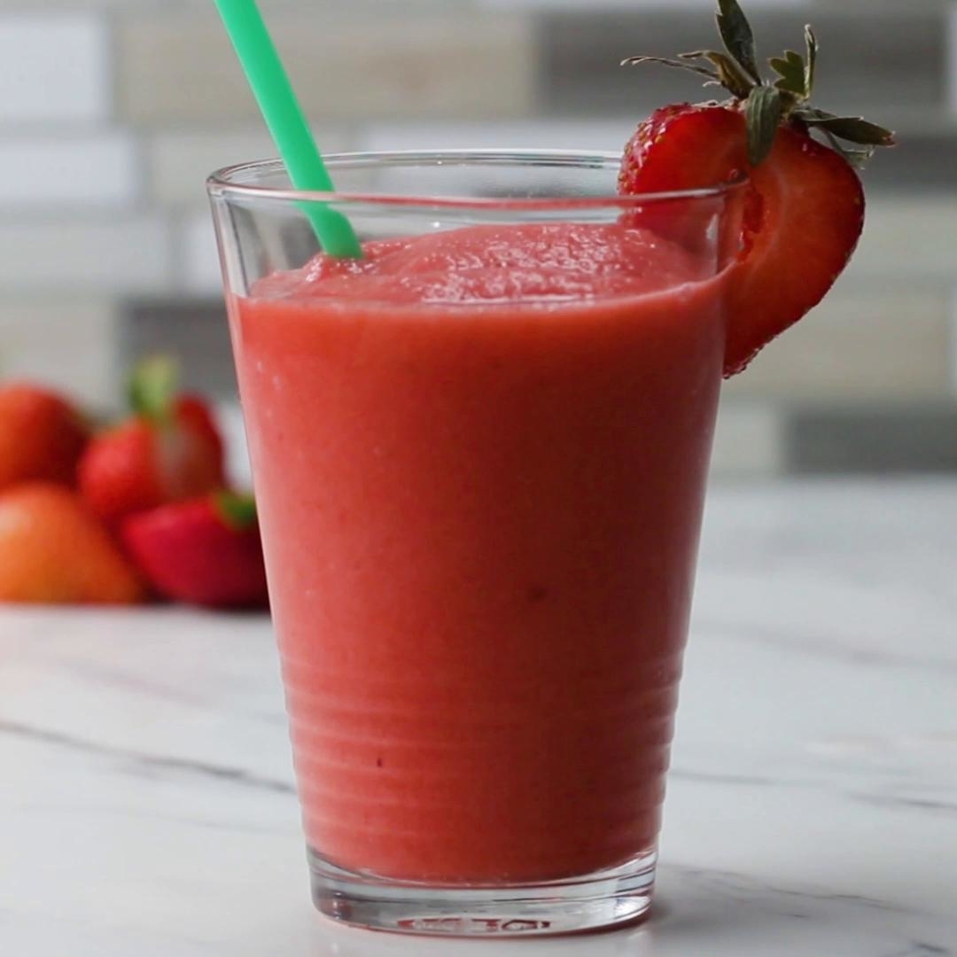 A strawberry smoothie in a glass with a green straw, garnished with a sliced strawberry