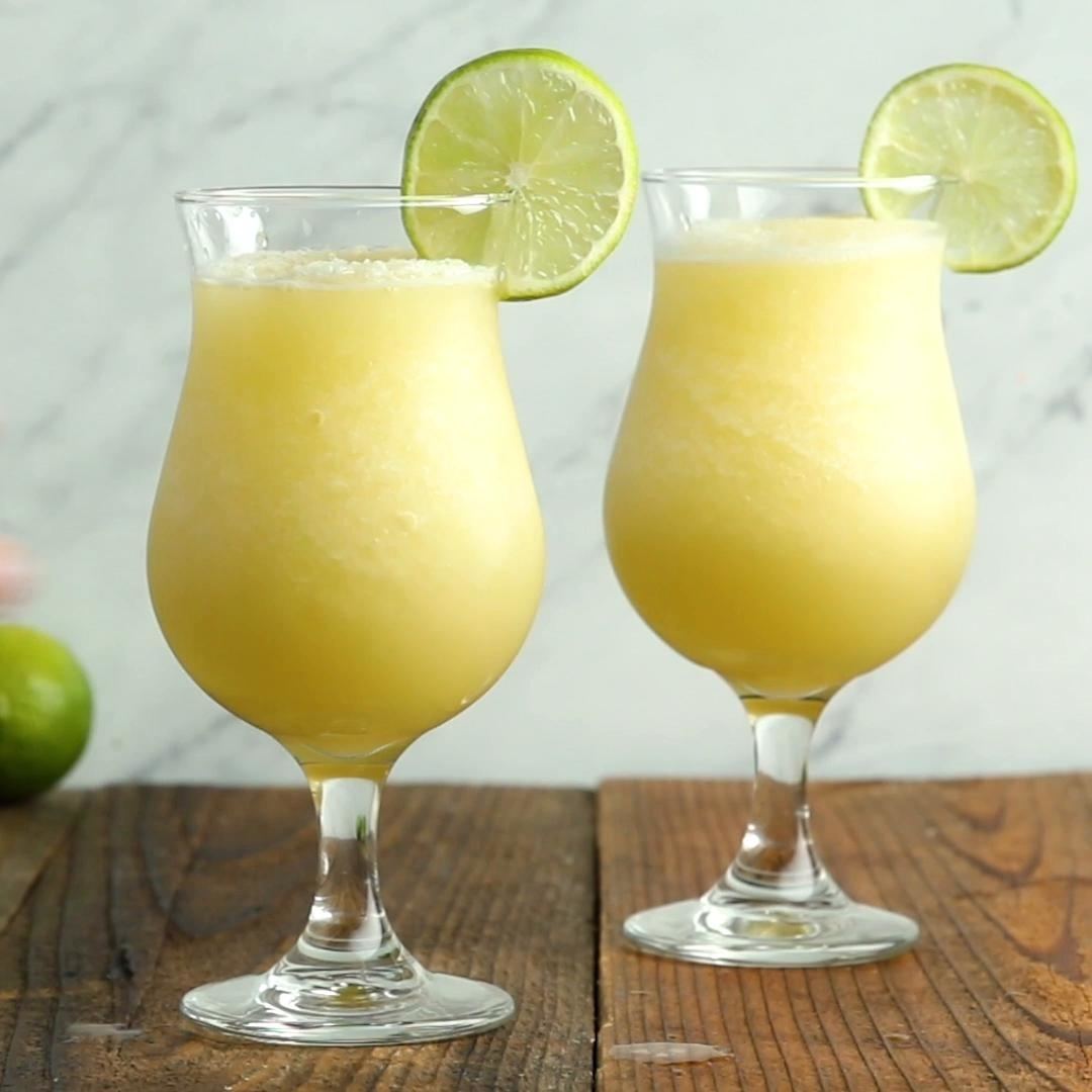 Two frozen margaritas in tall glasses garnished with lime slices sit on a wooden table, ready to be enjoyed