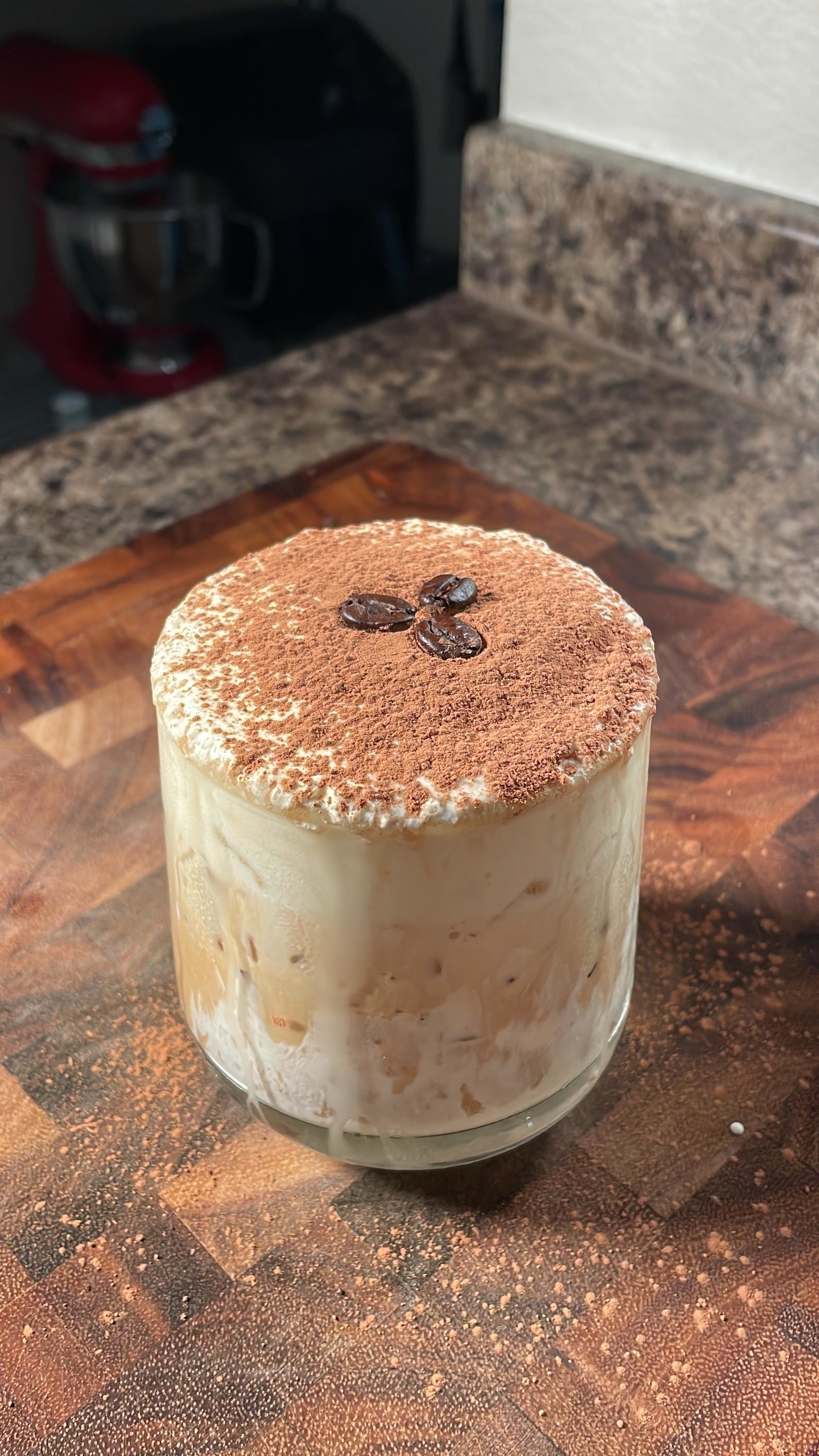 A creamy, iced coffee dessert in a glass, topped with cocoa powder and coffee beans, sits on a wooden cutting board