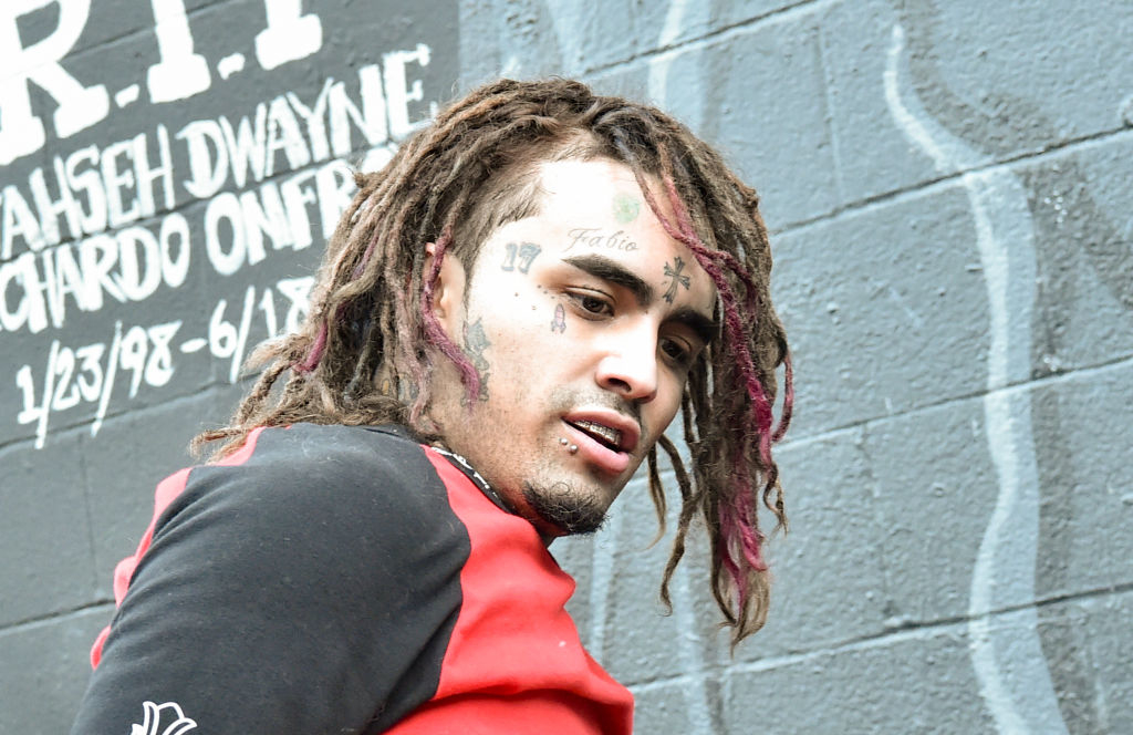 Lil Pump standing against a graffiti-covered wall, wearing a = shirt