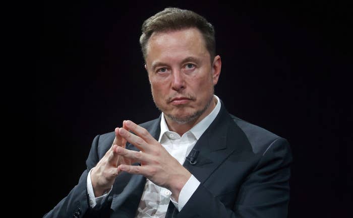 Elon Musk is seated, wearing a dark suit and light dress shirt, with hands clasped and an intense expression on his face