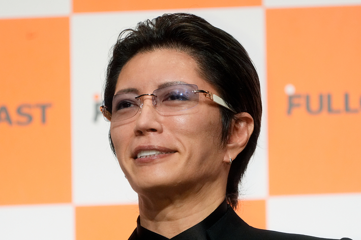 Gacktさんが、フルキャスト社のロゴが入った背景の前で、フォーマルな黒い衣装を着用している。