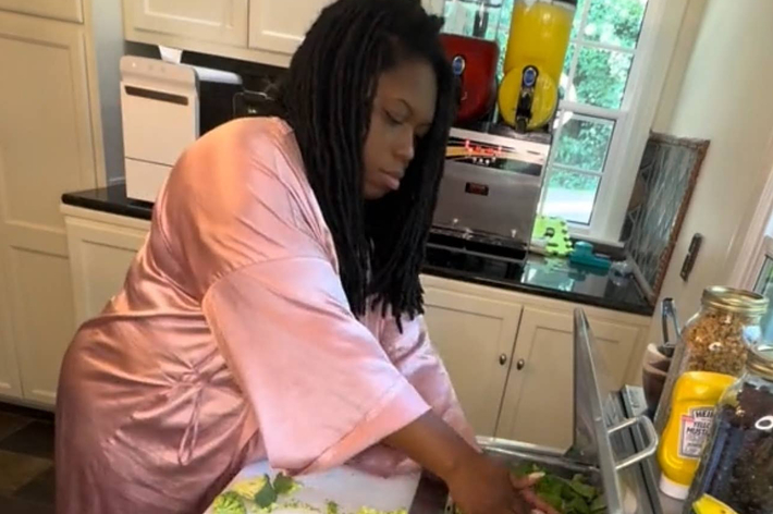 女性がキッチンで調理している。ドレッドヘアでピンクのローブを着ている。