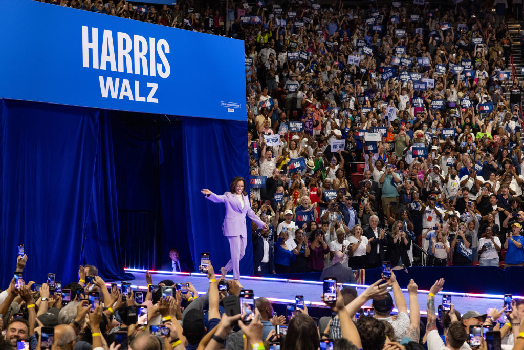 Kamala Harris walks onto a stage with people holding signs and phones. A blue banner reads, Harris Walz