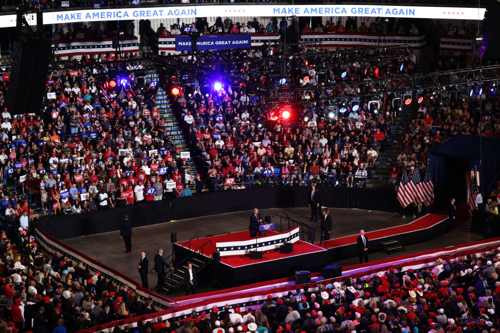 Rally with a crowd filling an arena. A stage with "Make America Great Again" signs is at the center