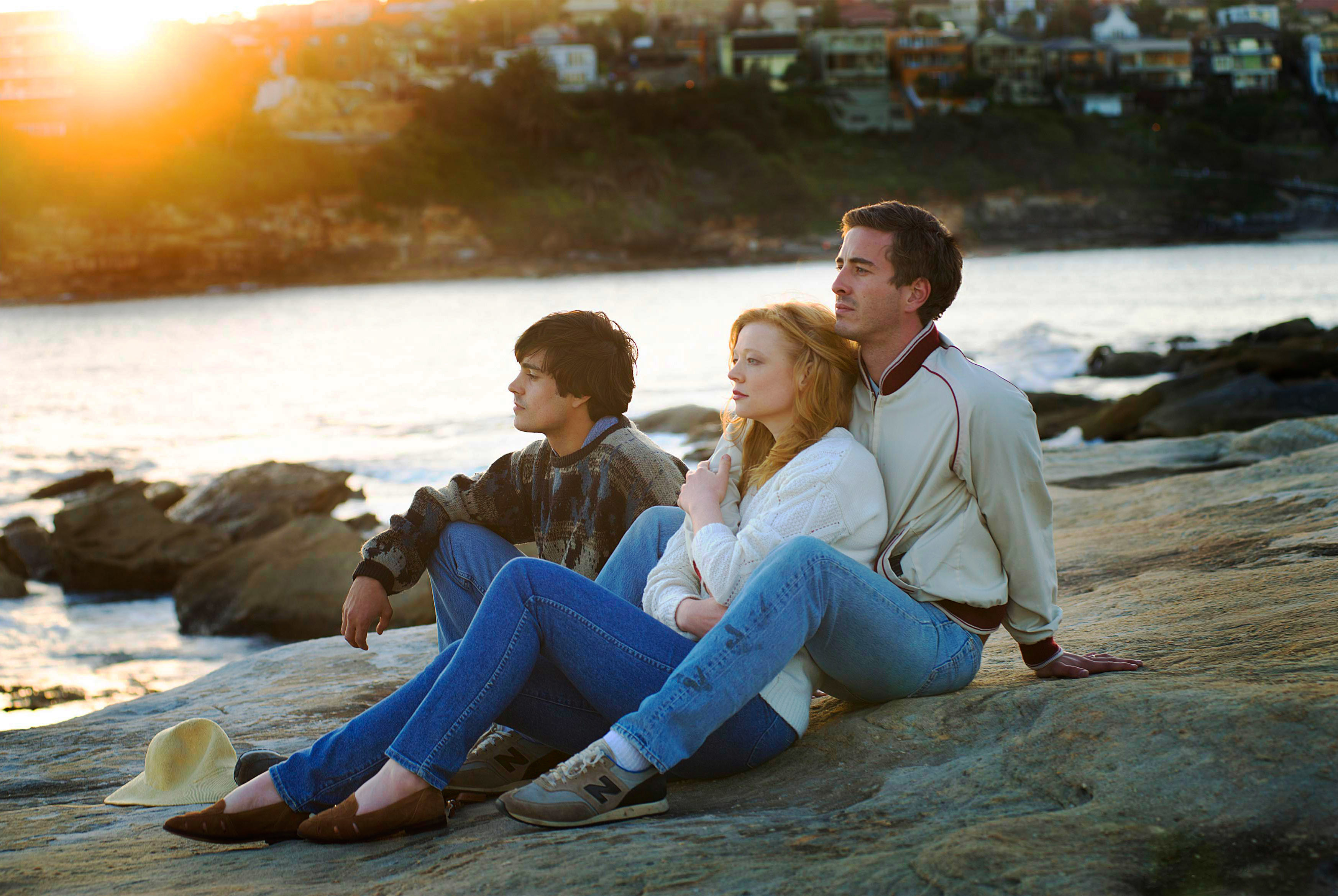 A family sits on rocky terrain by the sea during sunset: a teenage boy in a plaid shirt, a woman in a white sweater and jeans, and a man in a jacket and jeans