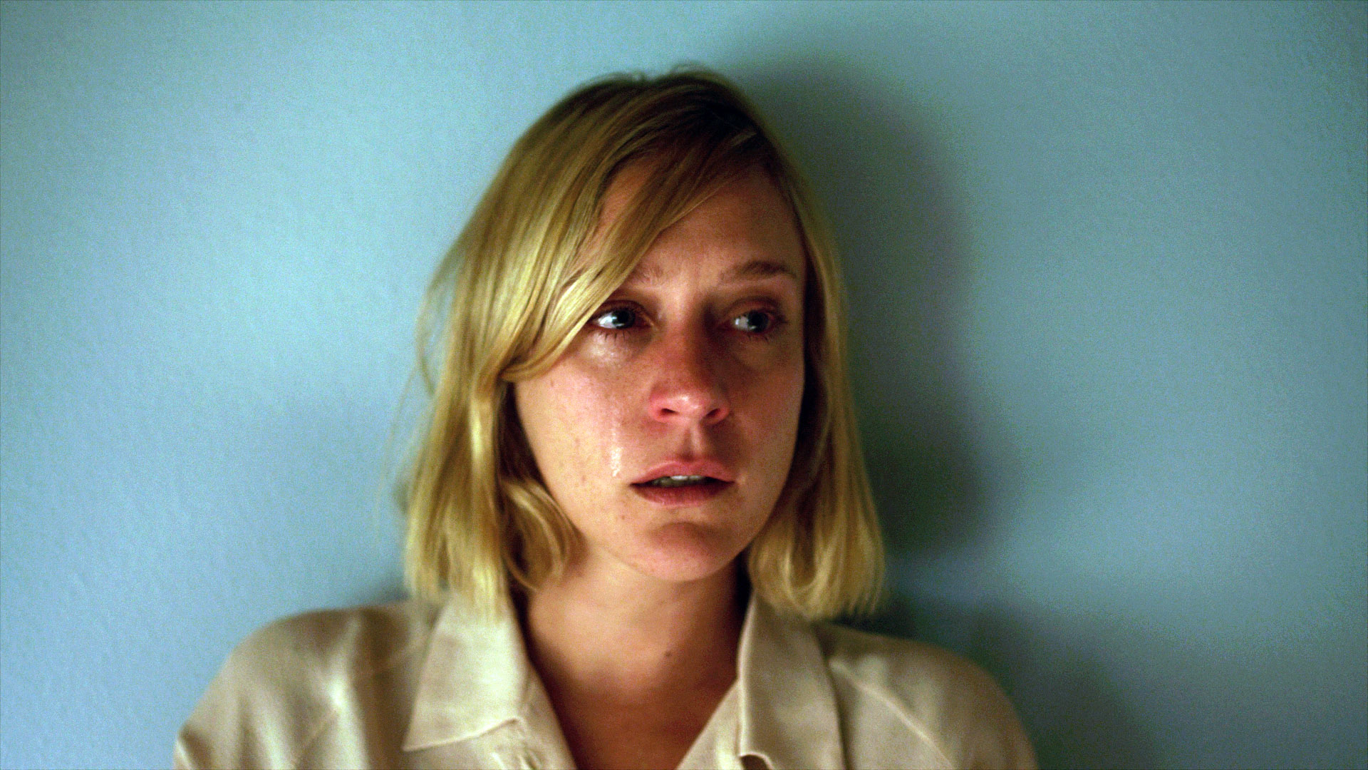 A woman with blonde hair and a distressed expression, wearing a light-colored shirt, stands against a plain background, appearing emotional