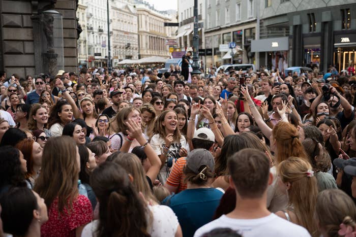 Crowd of people gathers on a city street, smiling and taking photos. Buildings and shops are visible in the background