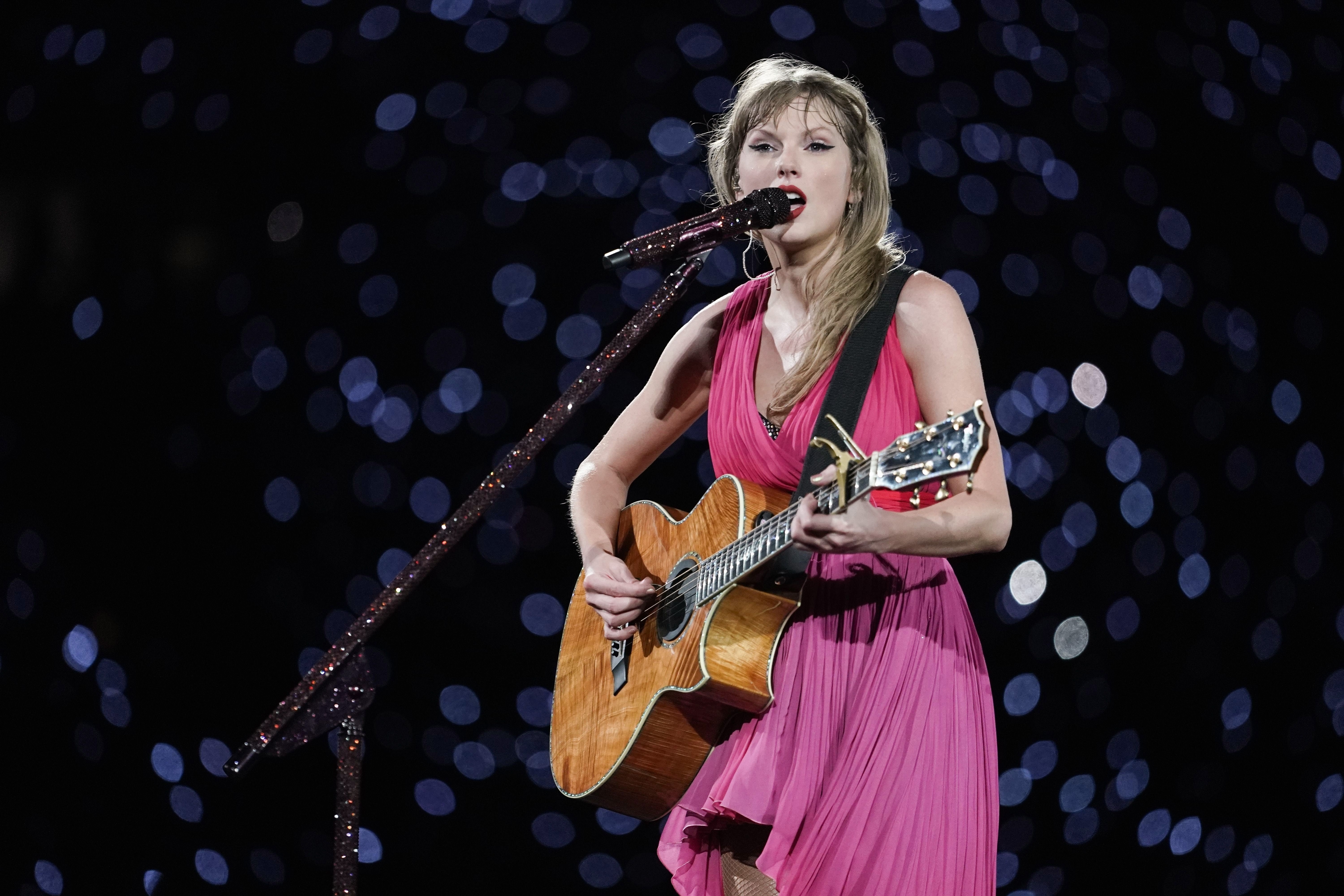 Taylor Swift performs on stage wearing a sleeveless, flowing dress, playing an acoustic guitar against a backdrop of out-of-focus lights